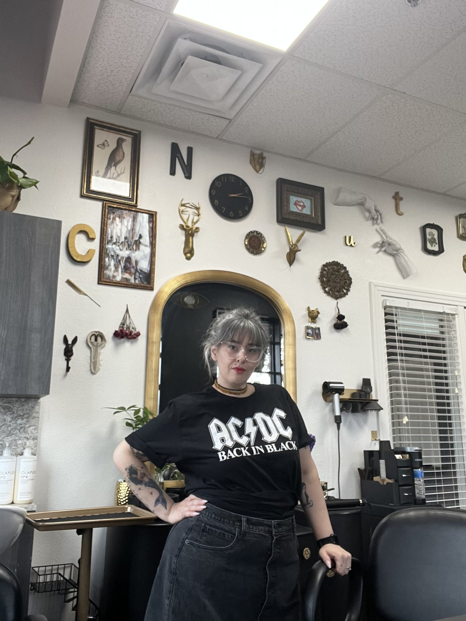Airika standing inside her studio with a wall decorated with various frames, letters, and decorative items. She is wearing a black AC/DC t-shirt, black jeans, glasses, and has tattoos on her left arm.